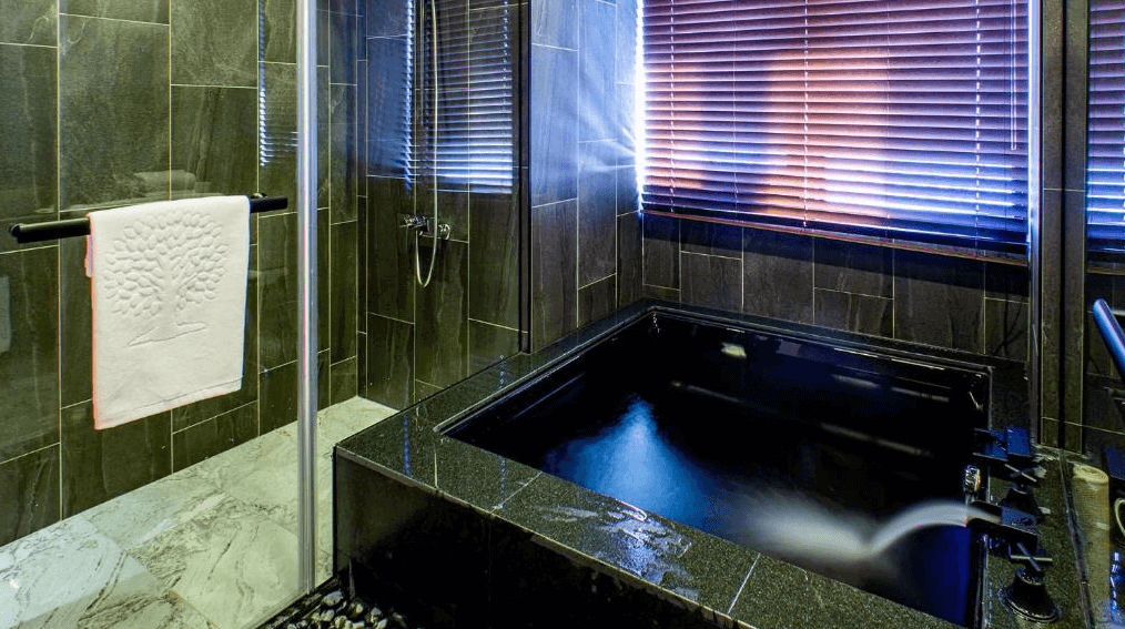 "台北小巨蛋住宿"甲山林湯旅的浴室圖片，內部配有一個黑色的大理石浴缸，浴缸周圍採用了深色瓷磚，營造出私密且高雅的氛圍。窗戶裝有木質百葉窗，能夠調節光線，提供更為私密的沐浴體驗。浴缸旁還設有淋浴區，設計風格現代且極具質感。