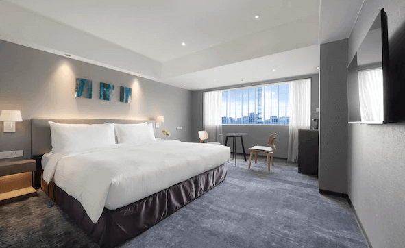 【台北小巨蛋住宿】茹曦酒店的酒店客房，房間配有一張大床，床鋪整潔，白色床單顯得乾淨明亮。房間以灰色調為主，裝飾簡約，並有幾幅抽象畫作點綴在床頭牆上。窗外可以看到城市景觀，光線充足，整體氛圍非常舒適和放鬆。