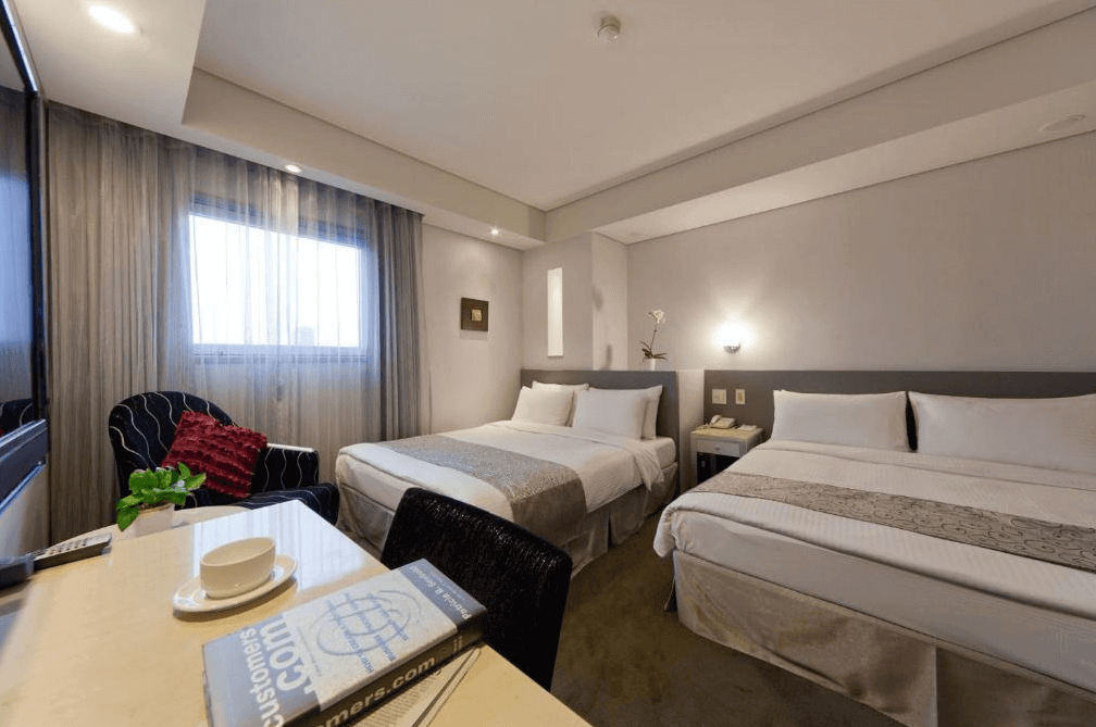 華帥海景飯店一間雙床房的內部照片，房間內有兩張雙人床，床頭背景牆為淺灰色，房間內還擺放了一張黑色扶手椅和小桌子，旁邊的窗戶掛著透明的紗簾，整體氛圍柔和舒適。