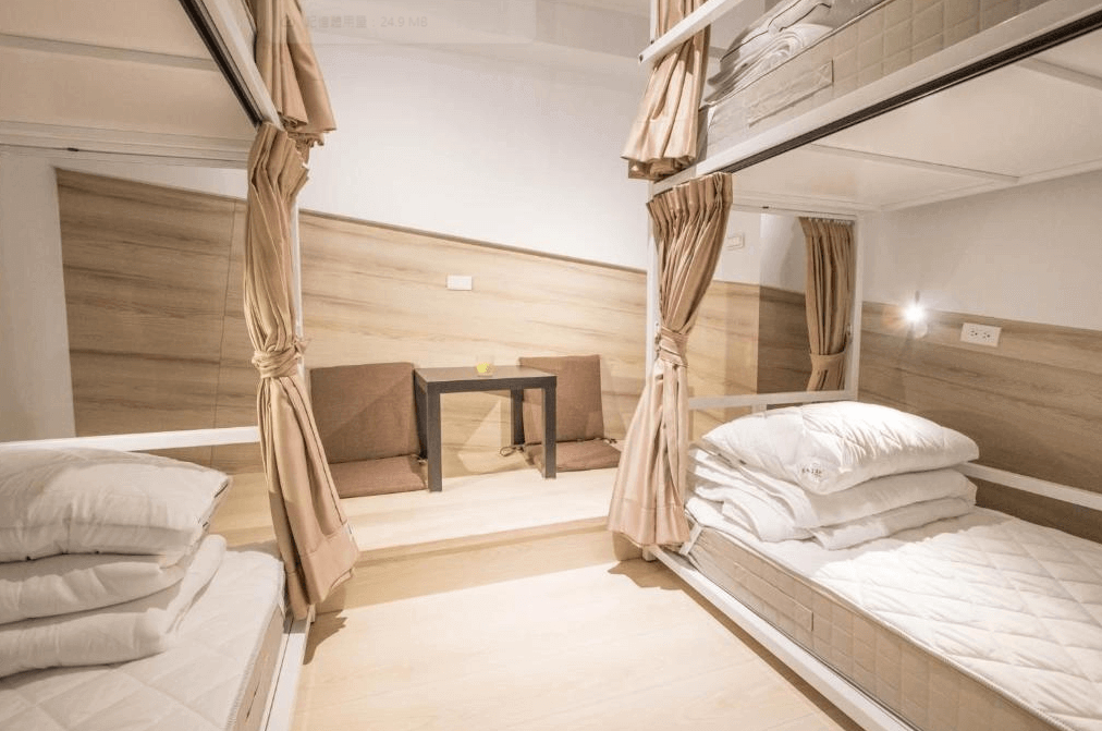 一間宿舍式房間的內部照片，房間內有上下鋪的床位，每個床位都配有布簾以提供隱私。牆角放置了兩個坐墊和一張小茶几，房間設計簡約且舒適，採用淺色木質材料。