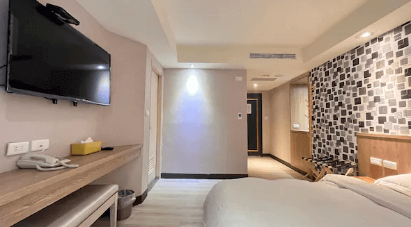 金華大飯店房間內的照片，展示了雙人床、牆上的平板電視以及設有桌椅的工作區域，牆壁上方仍然使用黑白方塊圖案的壁紙。房間設計簡約，色調溫暖，空間布置合理。