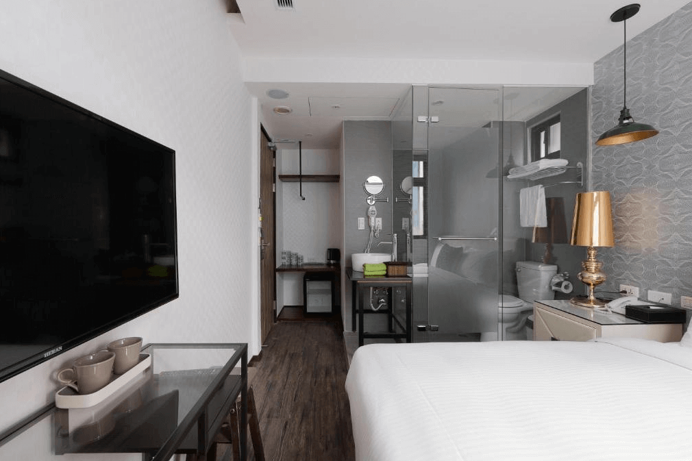 一間設計現代的客房內部照片，房間內有一張雙人床，床旁邊設有一個帶玻璃牆的浴室，浴室內設有洗手台和馬桶。牆壁上掛有一盞金色的桌燈，整個房間色調以白色和灰色為主，簡潔而現代。
