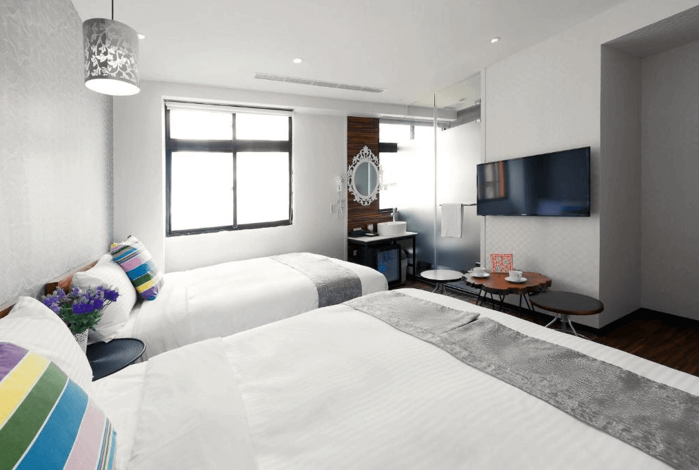 雙床房的內部照片，房間內有兩張雙人床，牆上裝飾有彩色條紋抱枕和紫色花卉裝飾，房間內設有一個帶玻璃牆的浴室，牆上掛有一面大電視，整體設計明亮且溫馨。