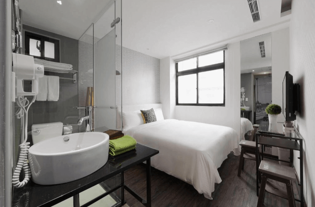 單人房的內部照片，房間內有一張雙人床，床旁邊是帶玻璃牆的浴室，內部配有洗手台和馬桶。房間設計簡約，白色的牆壁搭配深色地板，營造出舒適的氛圍。