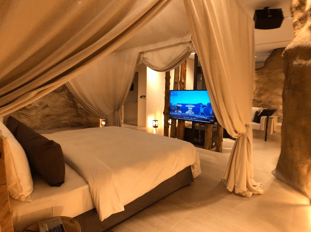 【台中情趣汽車旅館】鳥人創意旅店房間給人一種神秘的洞穴感，床四周圍繞著柔軟的帷幔，營造出私密且舒適的氛圍。電視被巧妙地嵌入在設計中，使得房間既實用又充滿創意。整體色調溫暖，讓人感到放鬆和愉悅。