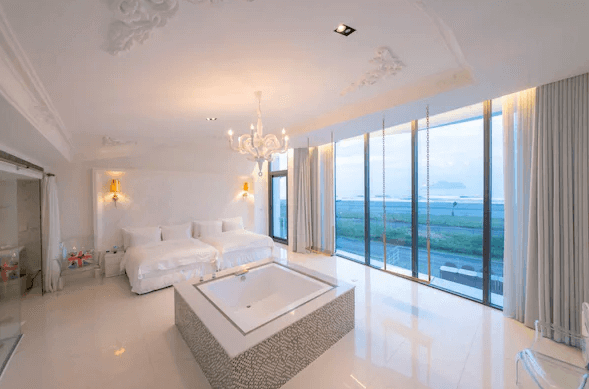 “頭城住宿”宜蘭PLAY Hotel飯店的一間奢華的雙床房，房間內有兩張大床，房間中央設有一個大型浴缸。房間的牆面和地板以白色為主，整體裝飾華麗而現代，天花板上有精美的雕花裝飾，並配有水晶吊燈。落地窗外可以欣賞到海景，光線明亮通透。