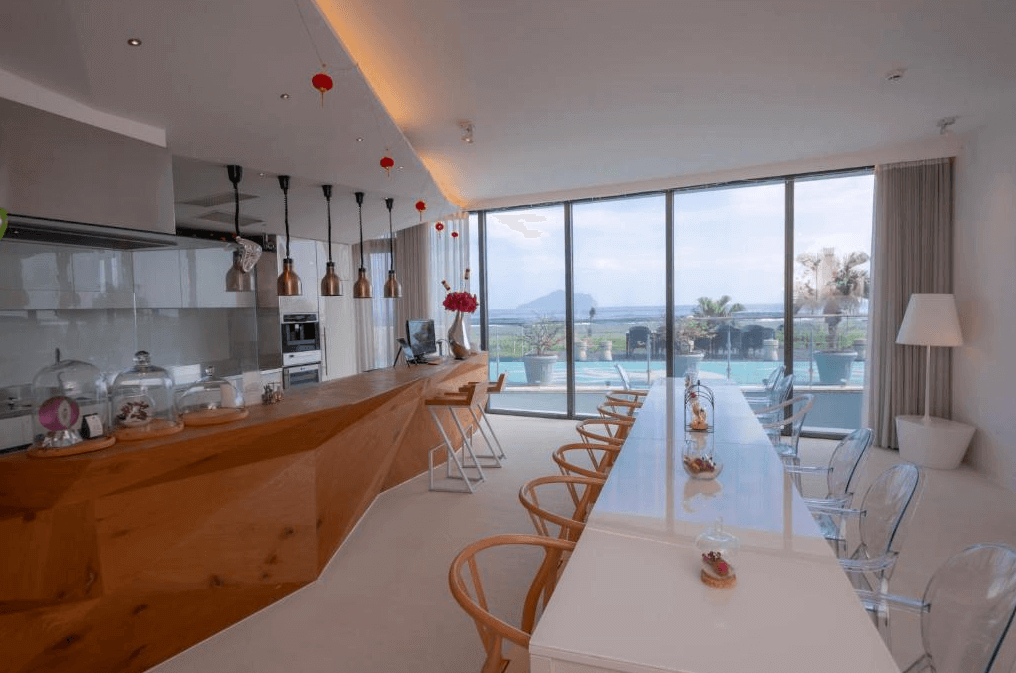 “頭城住宿”宜蘭PLAY Hotel飯店內的一個開放式餐廳，餐廳擺放著一張長桌，桌旁有透明椅子。木質的櫃台設計充滿幾何感，透過落地窗可以欣賞到泳池和外面的自然景色，光線充足，氛圍明亮愉快。