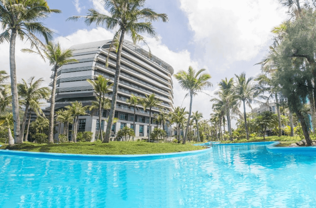 “頭城住宿”凱渡廣場酒店外部的景觀，建築四周環繞著椰子樹和其他熱帶植物。前方有一個藍色的泳池，水面閃耀著陽光，整體環境營造出熱帶度假的氛圍。