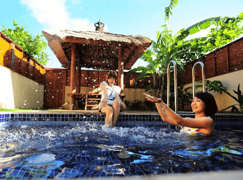 【墾丁寵物友善住宿】墾丁福華的戶外游泳池區域，水面上嬉戲著兩位女性。游泳池旁有熱帶植物圍繞，背景中可以看到茅草屋頂的涼亭，充滿濃濃的熱帶度假風情，給人放鬆愉快的感受。