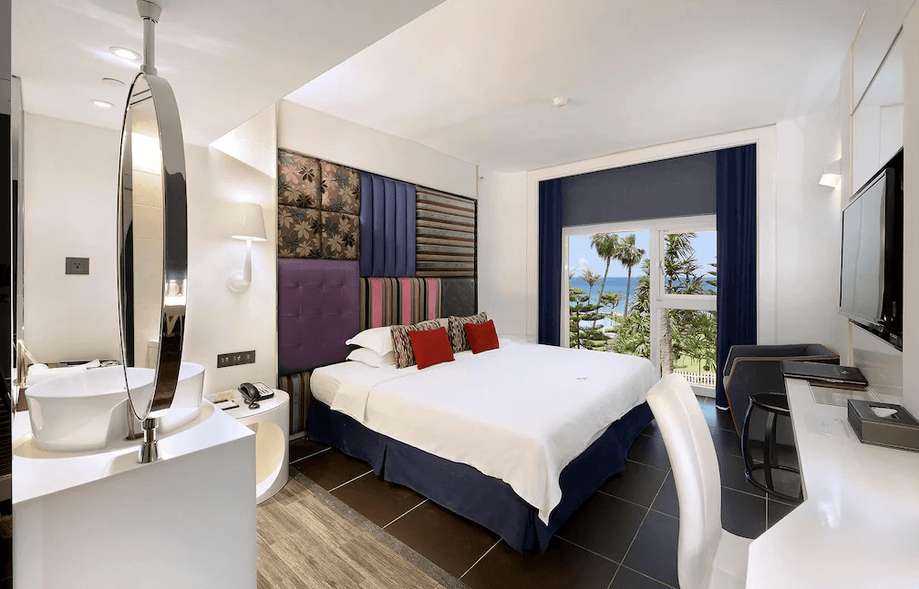 【墾丁寵物友善住宿】"墾丁海景住宿"夏都沙灘酒店的一間設計現代且時尚的飯店房間，內有一張大床，背景牆帶有獨特的裝飾圖案，窗戶外可看到海景和棕櫚樹，房內配有電視、辦公桌，以及靠近床頭的洗手台，房間色調以柔和的米白和深藍為主。