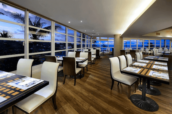 【墾丁寵物友善住宿】"墾丁海景住宿"夏都沙灘飯店餐廳的內部裝潢以現代簡約風格為主，餐桌排列整齊，白色和深棕色的座椅搭配木地板，窗戶外可以欣賞日落的美景，營造出寬敞且舒適的用餐氛圍。