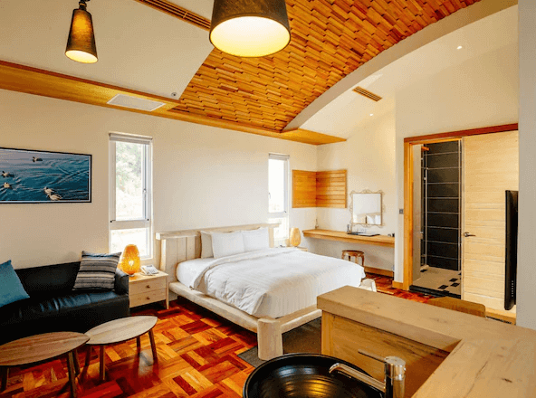 【墾丁海景住宿】"恆春住宿"海灣森林精品民宿的房間，木質天花板和牆面讓空間充滿自然感。房間內有一張大床，旁邊是黑色沙發和小茶几，整體色調溫暖，並配有木質傢俱，呈現出溫馨而舒適的氛圍。