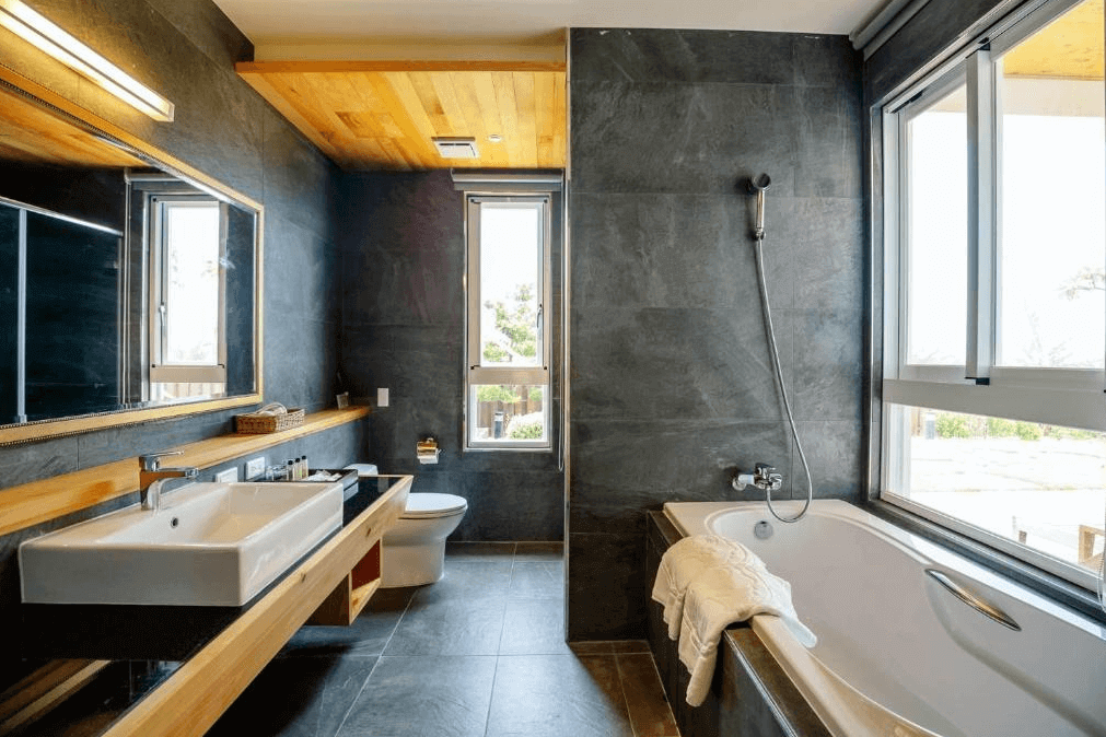 "恆春住宿"海灣森林精品民宿設計高雅的浴室，牆面以深灰色為主，搭配木質天花板。浴室內設有寬敞的浴缸和洗手台，旁邊的窗戶讓陽光透入，增添了空間的明亮感。