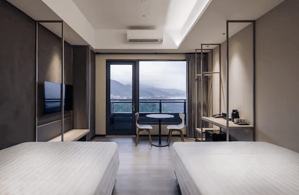 “頭城住宿”烏石港 OA HOTEL的一間雙床房，房內的兩張床對稱擺放，床頭有深色的背板設計。房間的落地窗外可以看到遠處的山景，房內設有小型圓桌和兩把椅子，適合享受窗外美景。