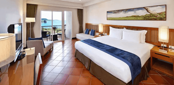 【墾丁寵物友善住宿】"墾丁海景住宿"福容大飯店的房間，設有一張大床，床鋪上有藍色的床尾巾，房間內的家具為木質設計，地板為紅棕色的瓷磚，窗外可以看到海景，並配有陽台。