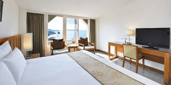 【墾丁寵物友善住宿】"墾丁海景住宿"福容大飯店的房間，設有一張大床，床前有書桌和椅子，窗戶外可見海景和藍天。房間內的裝潢以簡約木質風格為主，提供住客舒適的住宿環境。