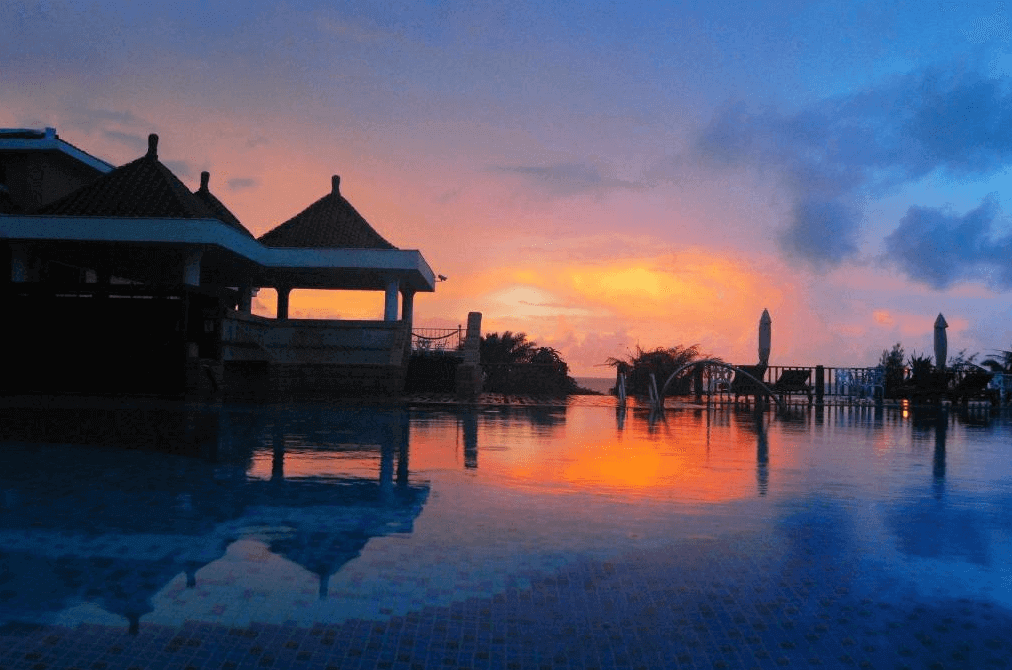 【墾丁寵物友善住宿】"墾丁海景住宿"福容大飯店夕陽下的泳池，池水倒映著橙黃色的晚霞，背景是飯店的傳統紅頂建築，營造出浪漫的度假景象。