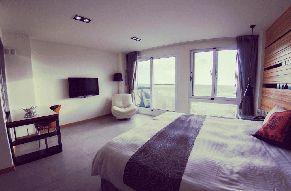 【墾丁寵物友善住宿】莎莎之拉夢民宿的面向海景的客房，擁有寬敞的雙人床，床單以灰色和白色為主，並有裝飾性圖案。房間內設有一個舒適的白色休閒椅和電視機，並且有一個通往陽台的大門窗，可以看到外面的海景。