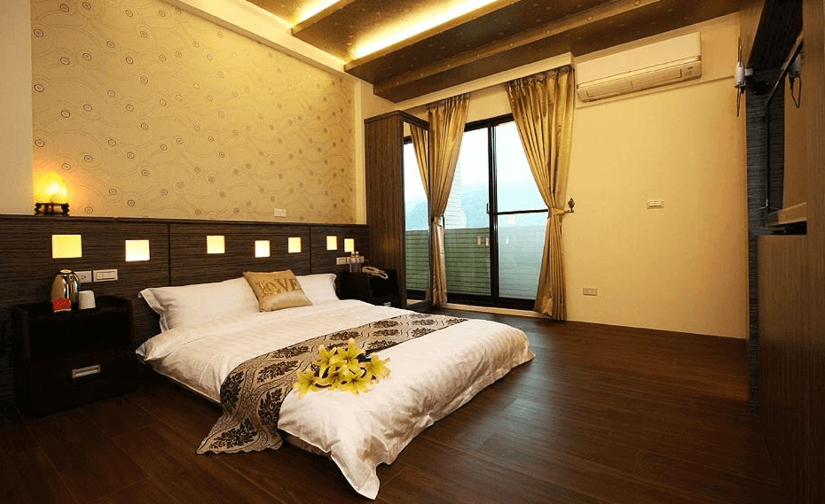 “頭城住宿”蘭博會館的一間溫馨的雙人房，房間內有一張大床，牆面設計以柔和的幾何圖案為主，並有溫暖的燈光點綴。房間的木地板增添了舒適感，落地窗旁邊有窗簾，窗外可以看到美麗的山景。