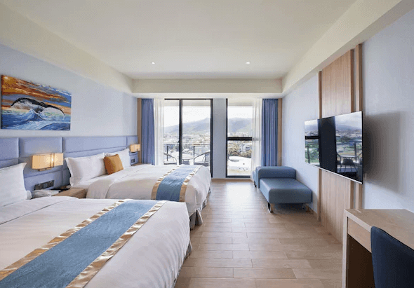 “頭城住宿”蘭陽烏石港海景酒店的一間帶有現代設計風格的雙床房，房間配備兩張大床，床鋪上有藍色和金色相間的床巾，牆上掛著一幅抽象風格的畫作。房間內的光線明亮，設有落地窗，可以看到外面的景觀。房間右側有一個大電視和藍色沙發椅。