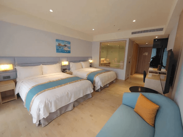 “頭城住宿”蘭陽烏石港海景酒店的一間雙床房，房間內兩張床整齊擺放，床上也鋪有藍色和金色相間的床巾。床旁邊有小型工作區，右側牆上有一扇透明玻璃窗，可看到浴室內部設施。房間內部裝修以淡雅色調為主，給人舒適感。