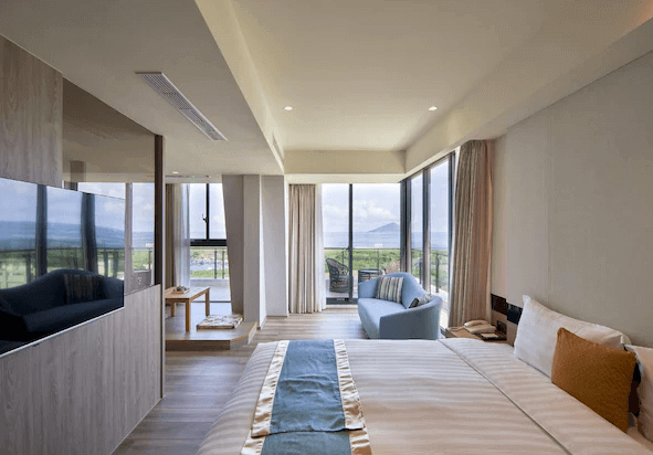 “頭城住宿”蘭陽烏石港海景酒店的一間大床房，房內擺放一張大床，床上有藍色和金色的床巾。房間四周採用全景落地窗設計，讓自然光線充分進入室內。外面可以看到綠意盎然的自然景觀和海景，還有一個小型休息區，配有沙發和茶几。