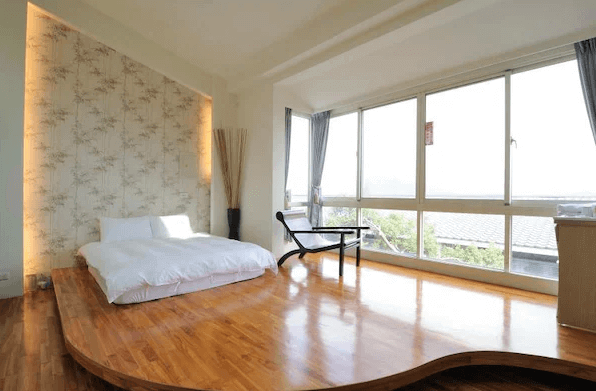 "三義住宿"三義幸福小築民宿的一間採光良好臥室，房間內設有一張簡約的日式風格低床，搭配木質地板，讓整個空間顯得明亮且寬敞。大面積的窗戶讓自然光充分進入，並能欣賞到窗外的景觀，營造出舒適放鬆的氛圍。