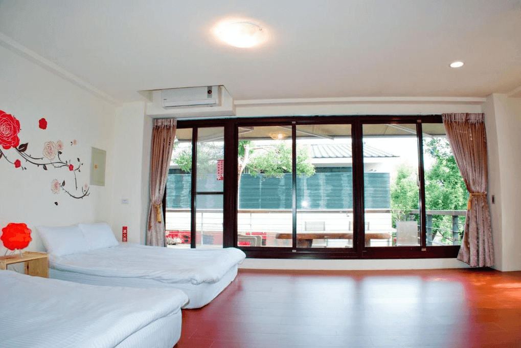 "三義住宿"三義幸福小築民宿的雙床房，房內有兩張雙人床，牆上有簡單的花卉壁貼裝飾，為房間增添了溫馨感。落地窗讓房間充滿自然光，並可通往外部露台，提供戶外休憩空間。