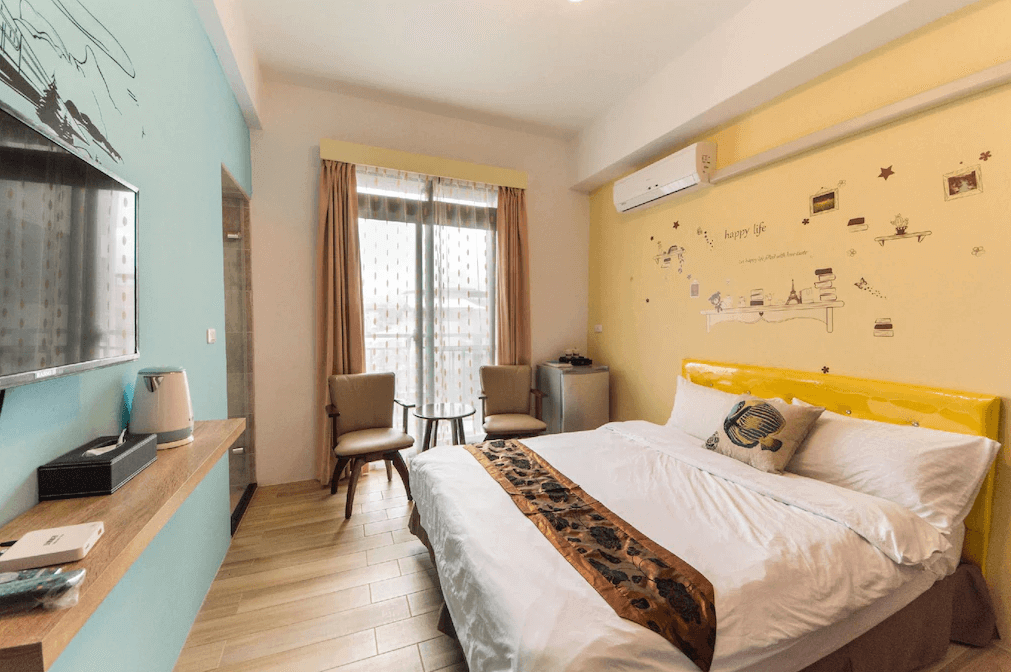 今古安民宿的淺藍色與黃色主題臥室，擺放著一張帶有黃色床頭板和裝飾枕的舒適雙人床。牆面分別採用淺藍色和黃色，並有可愛的牆面貼紙增添趣味。房間內配有一台大電視、一個茶壺，窗邊擺放了兩張椅子，營造出溫暖誘人的氛圍。