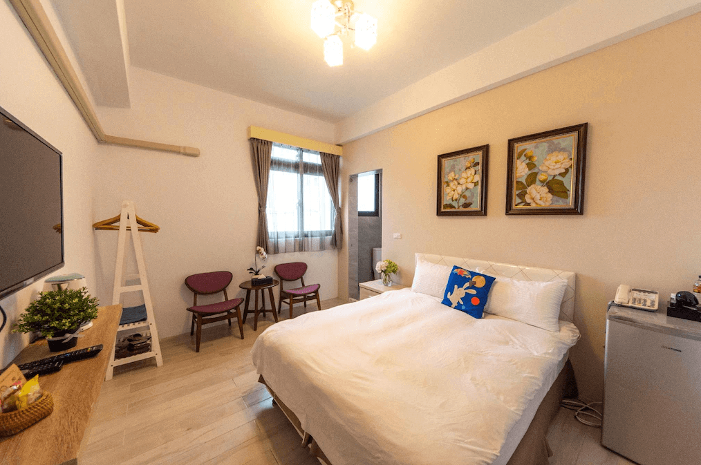 今古安民宿極簡風格的臥室，此房間設計風格簡約，以淡色調為主。雙人床上擺放著裝飾枕頭，牆上有藝術畫作點綴。房間內配有電視、小桌子和兩張椅子，並設有迷你冰箱等基本設備，呈現出簡約優雅的氛圍。