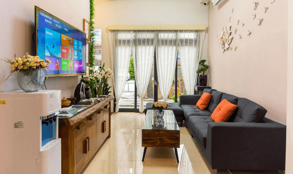 今古安民宿的客廳區域，一個風格時尚的客廳，擺放著一張配有橘色抱枕的灰色沙發。大電視掛在牆上，下面是木製的電視櫃。房間內還裝飾有植物和牆面藝術品，增添了溫馨歡迎的氣氛。