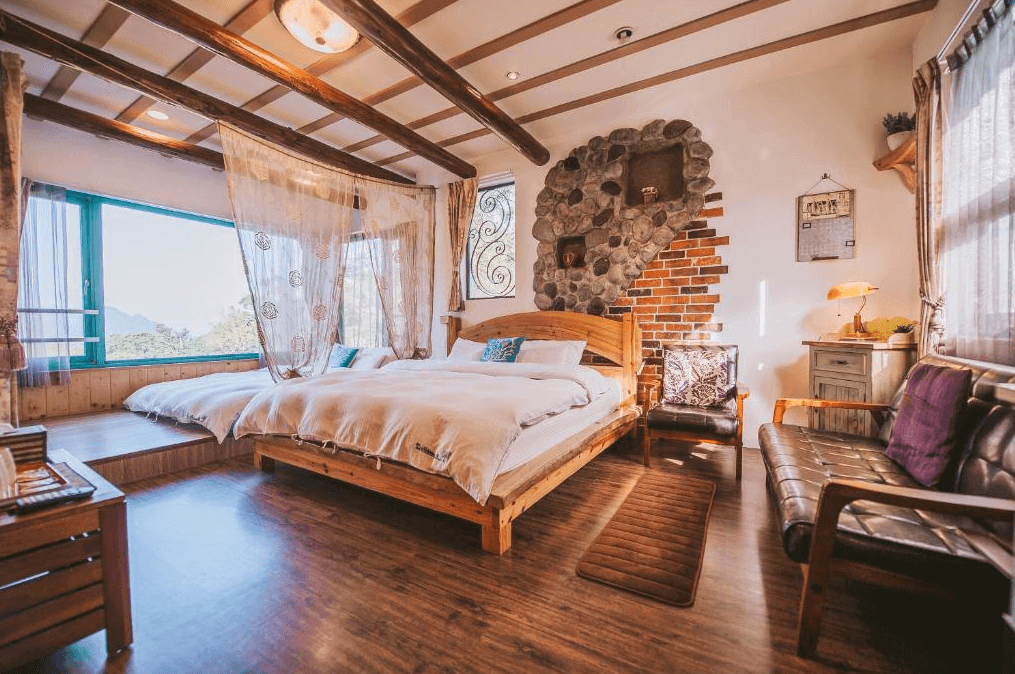 【南庄民宿】南庄橄欖樹咖啡民宿的臥室，房內有一張木質雙人床，床頭牆上裝飾著具有鄉村風格的石牆和紅磚。房間內光線充足，窗外可以欣賞到自然景觀，木質天花板上的橫樑增添了鄉村風的特色，整體給人一種溫暖而舒適的感覺。