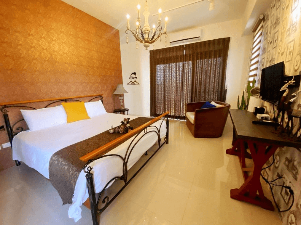 古月湖民宿的雙人房，牆面呈現金色紋理，搭配精緻的吊燈。房間內有一張金屬框架的雙人床，床上有白色床單和黃色抱枕。窗邊有落地窗，窗簾為深色系，旁邊還有一張舒適的椅子和木質書桌。