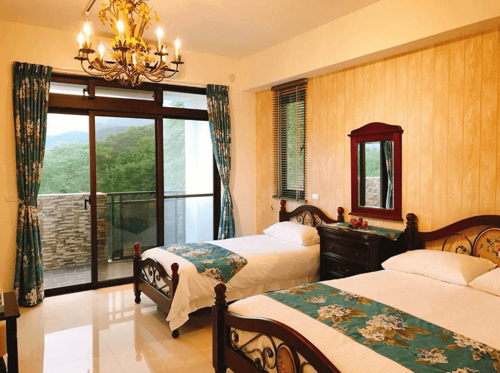 古月湖民宿的雙床房，擺設兩張古典風格的單人床，床上鋪有白色床單和花卉裝飾的床旗，吊燈為黃銅色風格。房間內有大面積落地窗，窗簾為綠色花卉圖案，窗外可見山林景色，營造出舒適的自然氛圍。