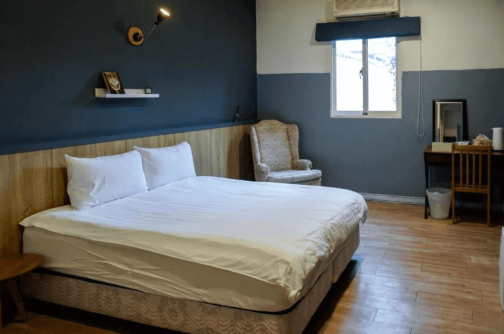 小和民宿的臥室，房內設有大床，牆壁是深藍與灰白色的雙色設計，房間內有一張舒適的扶手椅和木質梳妝桌，整體氛圍寧靜且舒適。