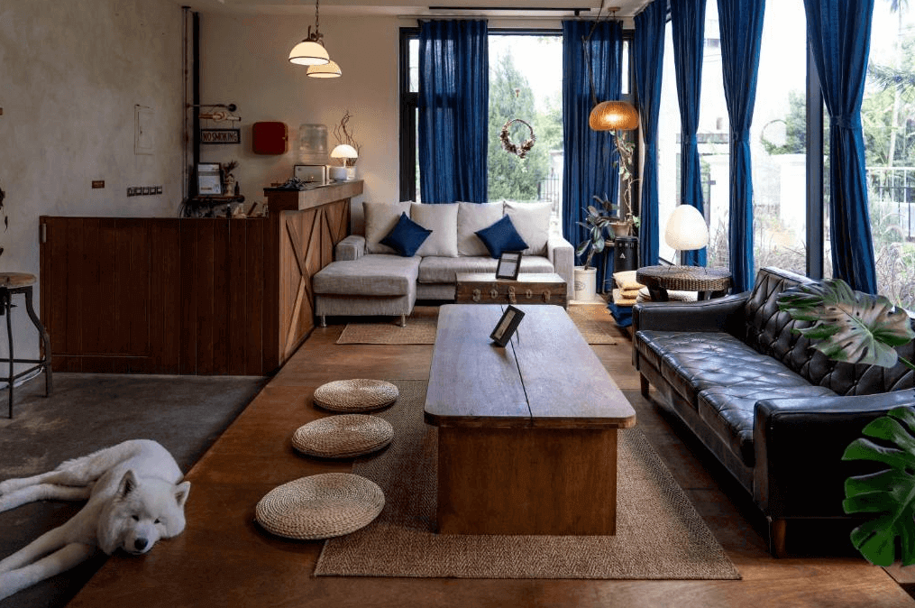 小和民宿的公共客廳區，內部擺設以木質和皮質家具為主，窗簾為深藍色，光線透過落地窗照亮整個空間，地板上鋪有編織地毯，旁邊還有一隻白色的狗慵懶地躺在地上，增添了一份家的溫馨感。