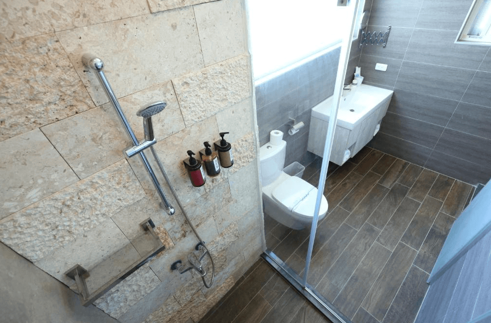 微藍海岸旅宿的浴室，採用質樸的石材和深色地板，搭配玻璃淋浴間，呈現出自然且高雅的風格。浴室設備齊全，設有洗手台、馬桶和壁掛式沐浴乳，為旅客提供方便的衛浴環境。