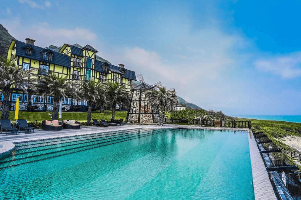 “花蓮寵物友善飯店”斯圖亞特海洋莊園民宿外觀及其戶外游泳池區域，建築風格為典型的歐洲莊園，明亮的黃色外牆與青翠的棕櫚樹相互映襯，戶外泳池的碧綠色水面閃耀著陽光，後方則是廣闊的海岸線，景色迷人。