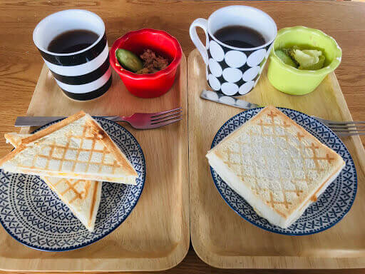 有人旅店的兩份早餐擺放在木盤中，包含方形三明治、咖啡和小菜。餐具與餐具的色彩對比顯得輕鬆愉快，這樣的擺盤讓人感受到清晨的愉悅氛圍，適合悠閒的早餐時光。