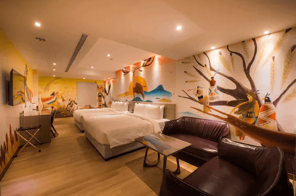 秝芯旅店駁二館的主題客房 這間房間充滿了鮮豔的藝術壁畫，牆面繪有原住民風格的圖案，兩張大床提供舒適的住宿空間。房間內還有深色皮沙發和木質小茶几，整體氛圍充滿創意與溫暖，適合家庭或朋友群體入住。