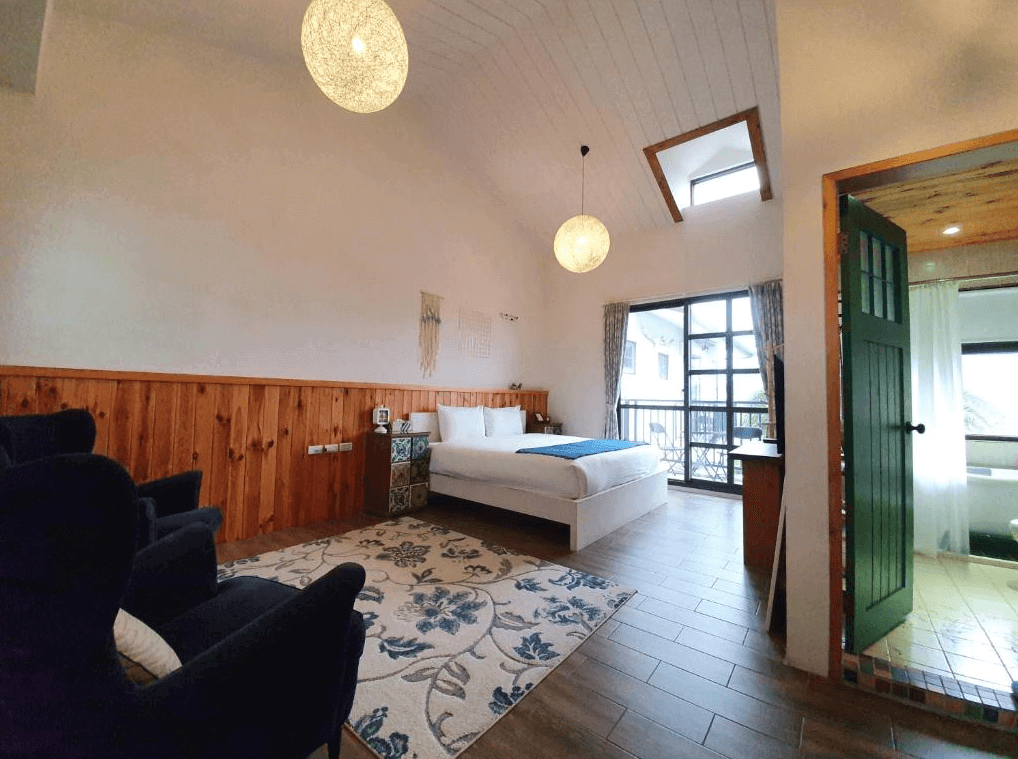 花蓮森林中民宿的溫馨舒適房間，設有木質牆壁裝飾與吊燈，白色床鋪上放有藍色裝飾毯，房間內有一張地毯以及黑色的沙發座椅，落地窗外有陽台和窗簾，整體風格自然且溫馨。