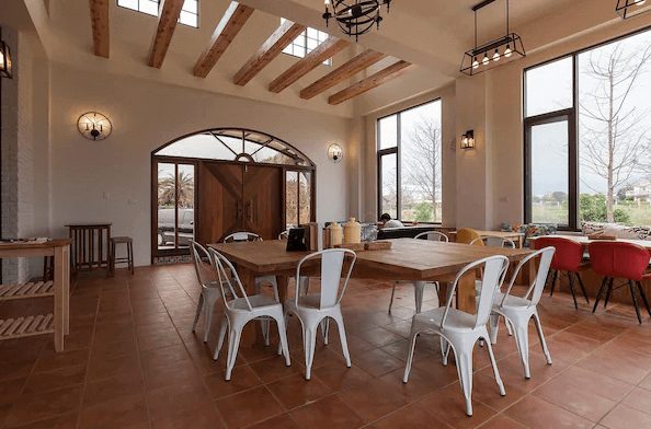 花蓮森林中民宿的公共用餐區，設有高挑天花板，木質桌椅整齊排列，大型落地窗讓自然光充滿整個空間，餐廳的裝潢簡約但富有質感，給人一種明亮而愜意的感覺。