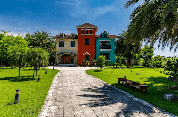 花蓮森林中民宿的外觀，建築顏色鮮豔，呈現多種顏色的外牆，前方是寬敞的綠色草坪和小徑，兩旁點綴著棕櫚樹，民宿被豐富的綠植圍繞，景色宜人。