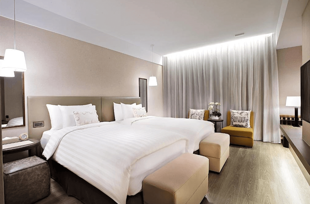 The GAYA Hotel潮渡假酒店豪華的雙床房,兩張床並排放置,房間採用溫暖的色調搭配簡約裝飾。床尾放置有柔軟的沙發椅與腳凳,營造出舒適放鬆的氛圍。牆上有精緻的吊燈,窗簾則是落地設計,增添高雅的感覺。