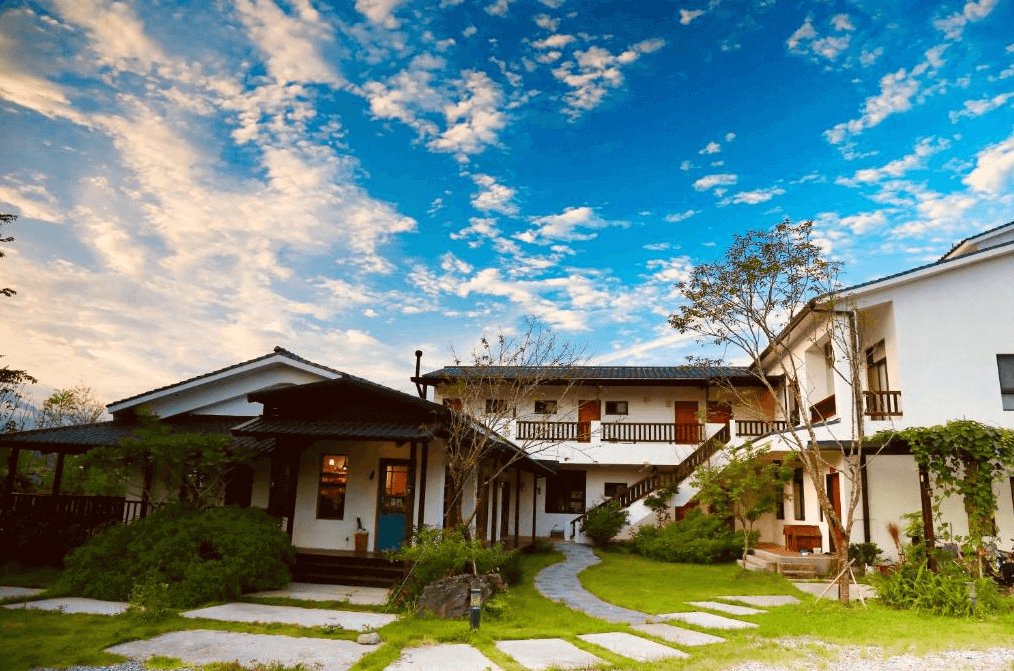 夏比安民宿的民宿外觀，建築以白色為主，配有木製陽台與綠意盎然的庭院小徑，藍天白雲映襯下，呈現出悠閒的田園風情。