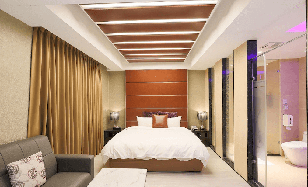 奇美國際時尚旅館的時尚客房，主牆面以橙色與金色元素點綴，搭配落地窗及柔軟的窗簾，房內有雙人床、沙發及透明玻璃隔間浴室，整體風格溫馨且現代。