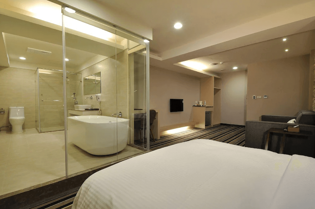 山水商務飯店的開放式套房，浴室使用透明玻璃牆，內有獨立浴缸與淋浴間，設計風格明亮簡潔。床鋪與沙發區一同位於房間內，空間感十足。