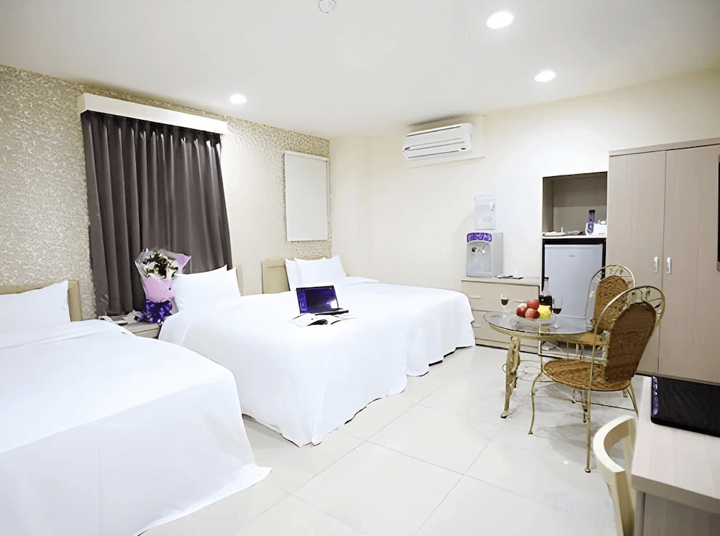 捷適商旅JS Hotel的客房，內部有三張白色床單覆蓋的雙人床，房間內設有小型玻璃桌和藤椅，旁邊有冰箱及飲水機。整體裝潢簡潔且舒適，窗戶有深色窗簾遮蔽。