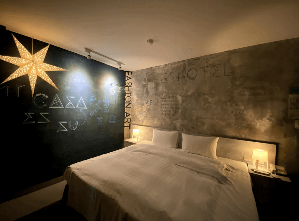 捷適商旅JS Hotel的客房，牆面採用工業風混凝土設計，並配有手繪藝術裝飾，包含星形吊燈及字母藝術元素，床上鋪有白色床單，兩側設有床頭燈。