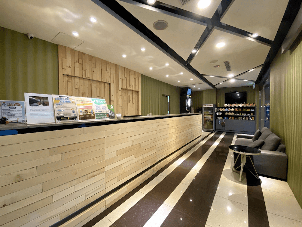 捷適商旅JS Hotel酒店接待大廳，前台以木質板材裝飾，背景牆配有綠色條紋設計，旁邊陳列著商品架和沙發區，整體環境明亮且現代。